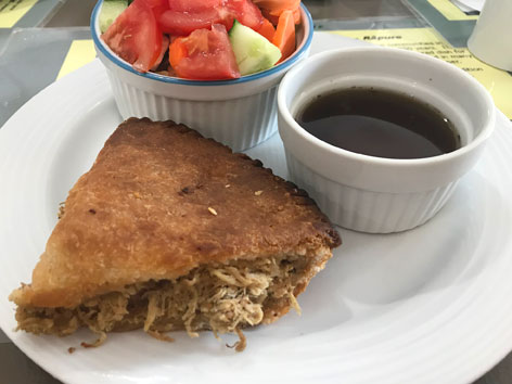 A triangular slice of Acadian meat pie with a side of beef jus and salad.  